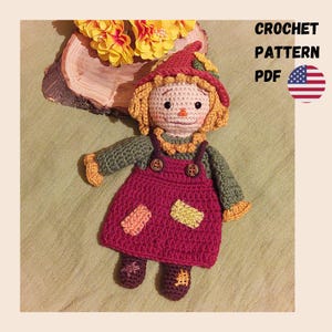May include: A crochet doll featuring a pointed hat, yellow hair, and a green long-sleeved shirt under a maroon overall dress with two brown buttons and two small rectangular patches. The text CROCHET PATTERN PDF is visible next to a US flag icon.