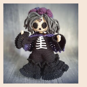May include: A handmade crochet doll with a spooky theme. The doll has gray hair, large black eyes, and a skeleton design on the front of its black dress. The doll's dress has a purple collar and accents. The doll is approximately 10 inches tall.