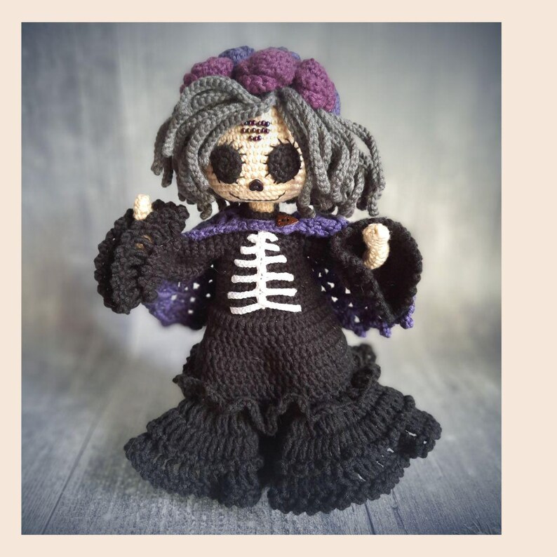 May include: A handmade crochet doll with a spooky theme. The doll has gray hair, large black eyes, and a skeleton design on the front of its black dress. The doll's dress has a purple collar and accents. The doll is approximately 10 inches tall.