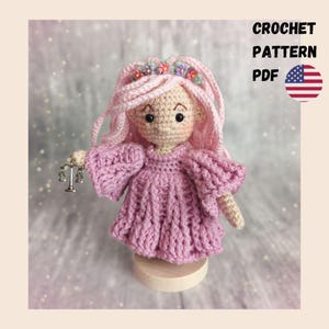 May include: A crocheted doll with light pink yarn hair adorned with small colorful flowers, wearing a textured light pink dress with wide sleeves. The doll holds a small silver-toned scale of justice charm. Text reads CROCHET PATTERN PDF.