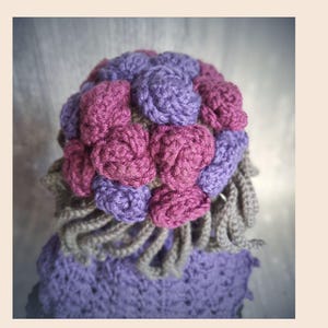 May include: A handmade crochet hat featuring a cluster of purple and magenta roses on top. The hat has a lavender base with a gray trim and a gray, curly fringe. The hat is a unique, handcrafted accessory.