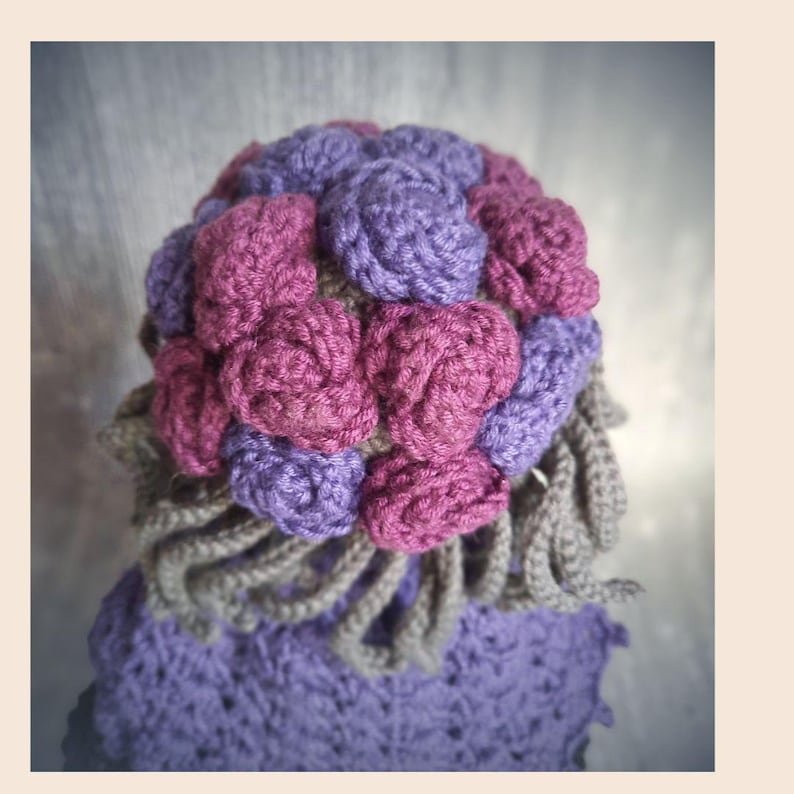 May include: A handmade crochet hat featuring a cluster of purple and magenta roses on top. The hat has a lavender base with a gray trim and a gray, curly fringe. The hat is a unique, handcrafted accessory.