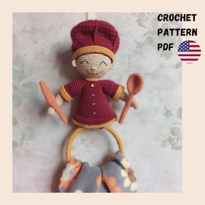 May include: A handmade crochet chef doll in a burgundy outfit with yellow trim, holding a spoon and knife. The doll has a smiling face and is attached to a towel ring with a patterned towel. Text on the right says "CROCHET PATTERN PDF" with an American flag.