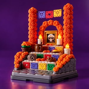 May include: A brick-built Dia de Muertos altar display featuring orange marigold flower columns, a central framed portrait, lit candles, and colorful papel picado banners. The tiered structure includes miniature food offerings and skulls on a stone-textured base, with the text KOZA DESIGN ALTAR DIA DE MUERTOS visible in the upper right corner.