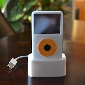 May include: A white digital music player with a black screen and an orange control wheel. It is docked in a white cradle, with a white USB cable attached. The device is on a dark brown wooden table.