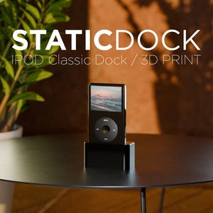 Op de afbeelding: Een zwart 3D-geprint iPod Classic-dock met het opschrift STATICDOCK IPOD CLASSIC DOCK / 3D PRINT. De rechthoekige houder bevat een zwarte iPod Classic met een kleurenscherm en een ronde clickwheel-interface.