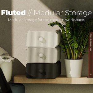 Puede incluir: Unidad de almacenamiento modular de tres cajones en blanco, gris y negro con diseño acanalado. La unidad tiene pomos redondos y está apilada sobre una estantería de madera. El texto "Fluted // Modular Storage" es visible. Una planta en maceta y libros también están en la imagen.