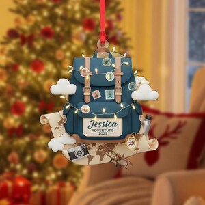 May include: A blue backpack Christmas ornament with a map, camera, and airplane. The ornament has the name "Jessica" and the text "Adventure 2025". It is decorated with string lights, clouds, and travel-themed stickers.