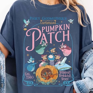May include: A navy blue t-shirt featuring a whimsical illustration from a fairy tale. The design includes the text "Fairy Godmother's Enchanted Pumpkin Patch" with cartoon characters and a pumpkin carriage. The shirt is a relaxed fit.
