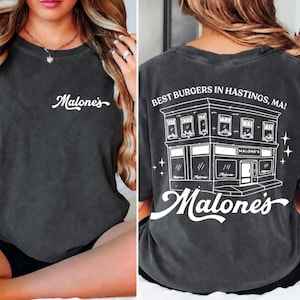 May include: A charcoal gray t-shirt featuring a small white script logo on the front chest. The back displays a large white line-art illustration of a building storefront with the text "BEST BURGERS IN HASTINGS, MA!" and "Malone's" below.