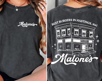 Puede incluir: Una camiseta gris carbón con un pequeño logotipo en letra cursiva blanca en el pecho. La parte posterior muestra una gran ilustración de líneas blancas de una fachada con el texto "BEST BURGERS IN HASTINGS, MA!" y "Malone's" debajo.
