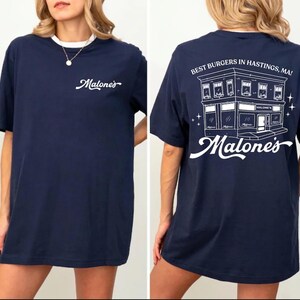 K&ouml;nnte beinhalten: Ein marineblaues T-Shirt mit einer wei&szlig;en Grafik auf der Vorderseite und dem Text Malone's. Die R&uuml;ckseite zeigt eine wei&szlig;e Strichzeichnung eines Geb&auml;udes mit dem Text Best Burgers In Hastings, MA! und dem Wort Malone's in Schreibschrift.