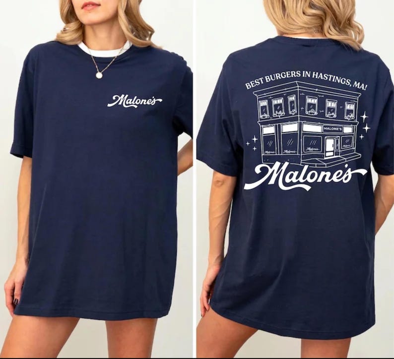 K&ouml;nnte beinhalten: Ein marineblaues T-Shirt mit einer wei&szlig;en Grafik auf der Vorderseite und dem Text Malone's. Die R&uuml;ckseite zeigt eine wei&szlig;e Strichzeichnung eines Geb&auml;udes mit dem Text Best Burgers In Hastings, MA! und dem Wort Malone's in Schreibschrift.