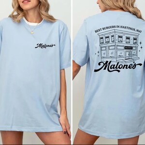 K&ouml;nnte beinhalten: Ein hellblaues T-Shirt mit dem schwarzen Schriftzug Malone's auf der linken Brust. Die R&uuml;ckseite zeigt eine schwarze Strichzeichnung eines Geb&auml;udes mit dem Text BEST BURGERS IN HASTINGS, MA! und dem Wort Malone's in Schreibschrift darunter.