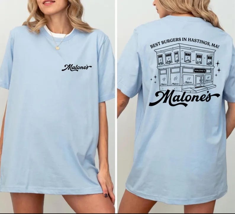 K&ouml;nnte beinhalten: Ein hellblaues T-Shirt mit dem schwarzen Schriftzug Malone's auf der linken Brust. Die R&uuml;ckseite zeigt eine schwarze Strichzeichnung eines Geb&auml;udes mit dem Text BEST BURGERS IN HASTINGS, MA! und dem Wort Malone's in Schreibschrift darunter.