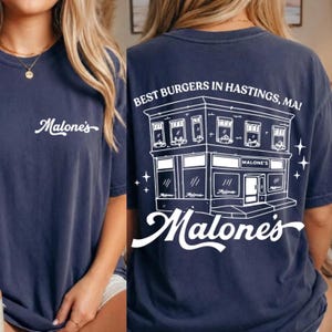 K&ouml;nnte beinhalten: Ein dunkelblaues T-Shirt mit einem wei&szlig;en Schriftzug auf der Brust. Die R&uuml;ckseite zeigt eine wei&szlig;e Strichzeichnung eines Geb&auml;udes mit dem Text "BEST BURGERS IN HASTINGS, MA!" und dem Schriftzug "Malone's" unter dem Geb&auml;ude.