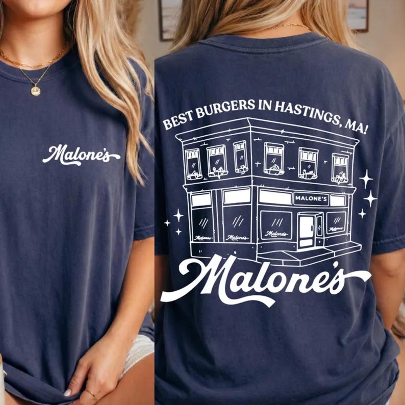 K&ouml;nnte beinhalten: Ein dunkelblaues T-Shirt mit einem wei&szlig;en Schriftzug auf der Brust. Die R&uuml;ckseite zeigt eine wei&szlig;e Strichzeichnung eines Geb&auml;udes mit dem Text "BEST BURGERS IN HASTINGS, MA!" und dem Schriftzug "Malone's" unter dem Geb&auml;ude.