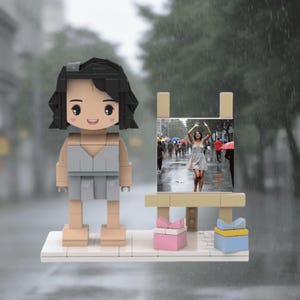 May include: A Lego-style figurine of a woman with black hair and a grey dress, standing beside a miniature easel displaying a photo of a woman dancing in the rain. The scene includes a white base, pink and blue gift boxes, and a rainy background.