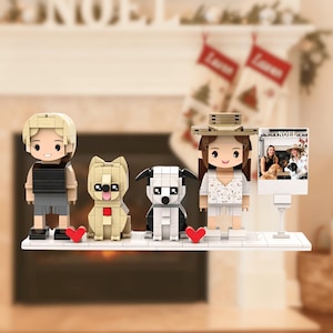 Puede incluir: Una estantería con figuras de bloques de dos personas, dos perros y una foto. Las figuras son de estilo caricatura, con los perros en beige y blanco y negro. Hay corazones rojos cerca de los perros. La foto muestra a dos mujeres y dos perros.