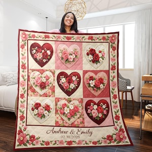 以下が含まれることがあります： 赤、ピンク、クリーム色のハート型の花柄が特徴のパーソナライズされたキルト。キルトは花の模様の縁取りがあり、「Andrew & Emily」の名前と日付「02.10.2025」が含まれています。