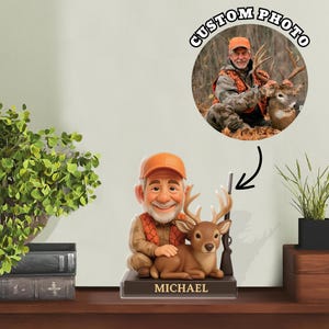 Puede incluir: Una figura cabezona personalizada de un cazador sonriente con barba, que lleva una gorra y un chaleco naranja, arrodillado junto a un ciervo marrón con astas y un rifle. La base es negra con el nombre MICHAEL impreso en letras doradas.