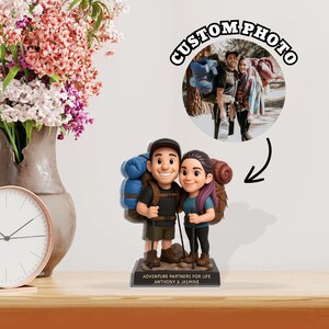 May include: A figurine of a smiling couple, each with a backpack and hiking poles, standing on a base that reads "ADVENTURE PARTNERS FOR LIFE ANTHONY & JASMINE." A circular photo above shows the couple hiking. The words "CUSTOM PHOTO" are above the photo.