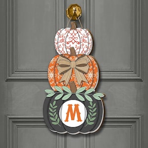 Könnte beinhalten: Ein Türhänger aus Holz mit Kürbis-Motiv, bestehend aus drei gestapelten Kürbissen. Der obere Kürbis ist weiß mit einem roten Muster, der mittlere ist orange mit einer Schleife und der untere ist schwarz mit einem weißen Kreis und dem Buchstaben "M".