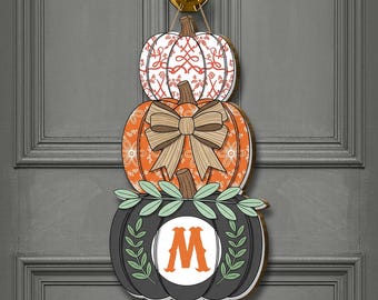 Puede incluir: Un colgador de puerta de madera con temática de calabaza con tres calabazas apiladas. La calabaza superior es blanca con un patrón rojo, la del medio es naranja con un lazo y la inferior es negra con un círculo blanco y la letra "M".