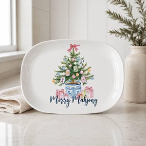 K&ouml;nnte beinhalten: Eine ovale wei&szlig;e Keramikplatte mit einer Aquarellillustration eines Weihnachtsbaums in einem blau-wei&szlig;en Topf, geschm&uuml;ckt mit Mahjong-Steinen und Kugeln, mit Geschenken am Fu&szlig; und dem Schriftzug Merry Mahjong.