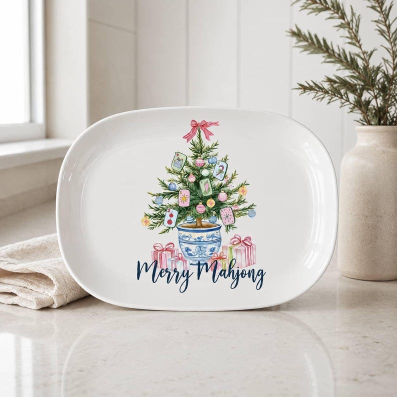 K&ouml;nnte beinhalten: Eine ovale wei&szlig;e Keramikplatte mit einer Aquarellillustration eines Weihnachtsbaums in einem blau-wei&szlig;en Topf, geschm&uuml;ckt mit Mahjong-Steinen und Kugeln, mit Geschenken am Fu&szlig; und dem Schriftzug Merry Mahjong.