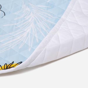 Puede incluir: Una alfombrilla de tela acolchada con fondo azul claro, patrones de hojas de palma dibujadas en blanco y detalles florales amarillos. La alfombrilla tiene un borde blanco acolchado y una textura de costuras en forma de diamante.