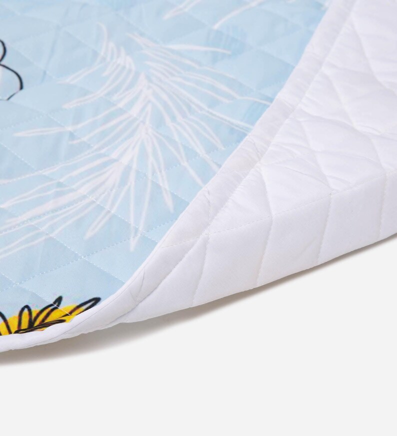 Puede incluir: Una alfombrilla de tela acolchada con fondo azul claro, patrones de hojas de palma dibujadas en blanco y detalles florales amarillos. La alfombrilla tiene un borde blanco acolchado y una textura de costuras en forma de diamante.