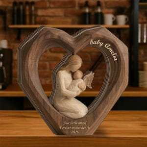 May include: A wooden heart-shaped frame with a carved mother and child angel figurine. The frame has the words "baby Amelia" and "Our little angel Forever in our hearts 2026" engraved on it. The frame is made of a dark brown wood.