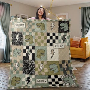 May include: A quilted blanket featuring a patchwork pattern of sage green and white checkered squares, black and white scribbled squares, and black and white checkerboard squares. The design includes dinosaur heads, lightning bolts, and the name WILLIAM.