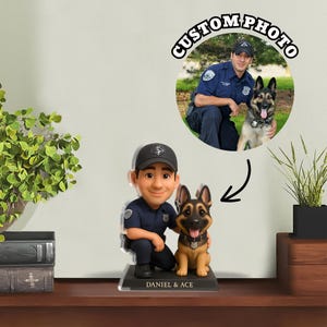 Può includere: Una statuetta bobblehead di un agente di polizia in ginocchio accanto a un cane pastore tedesco. La statuetta è su una base nera con i nomi "DANIEL & ACE". Sullo sfondo c'è una foto di un agente di polizia e di un cane con la scritta "CUSTOM PHOTO".