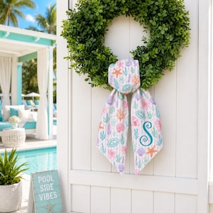 May include: A green boxwood wreath with a decorative scarf featuring a seashell pattern in pink, orange, and blue. The scarf is tied to the wreath and has a teal letter "S" on it. The wreath hangs on a white door.