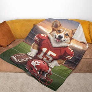 Puede incluir: Una manta con un perro corgi sonriente que lleva una camiseta de f&uacute;tbol roja y blanca con el n&uacute;mero 15. Un bal&oacute;n de f&uacute;tbol y un casco rojo con una huella de pata tambi&eacute;n est&aacute;n en la manta, que tiene un dise&ntilde;o de campo de f&uacute;tbol y luces de estadio.
