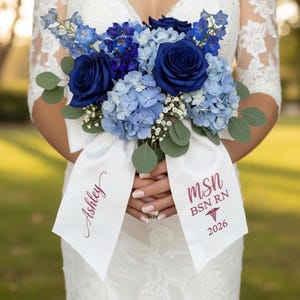 May include: A bouquet of blue roses, hydrangeas, and greenery, held together with a white ribbon. The ribbon is printed with the name "Ashley" and the text "MSN BSN RN 2026". The flowers are in shades of blue.