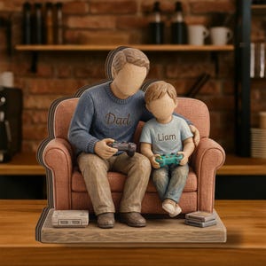 May include: A figurine of two figures sitting on a textured peach-colored sofa. The larger figure wears a blue sweater with the word Dad, and the smaller figure wears a light blue shirt with the name Liam. Both hold game controllers.