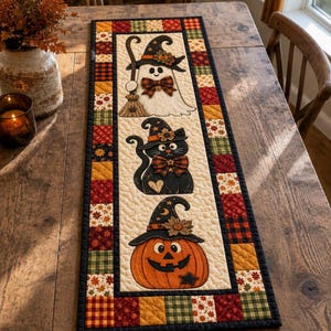 May include: A rectangular quilted table runner featuring a cream-coloured centre panel with three appliqued figures: a white ghost with a broom, a black cat, and an orange jack-o'-lantern, each wearing a witch hat. The border is a patchwork of autumn-themed fabrics.