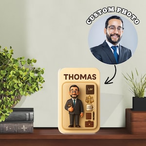Puede incluir: Un accesorio de escritorio personalizado con temática de abogado que presenta una figura de dibujos animados, el nombre "THOMAS" y una foto personalizada. El diseño incluye iconos relacionados con la ley y un maletín, todo en 3D. El fondo es beige neutro.