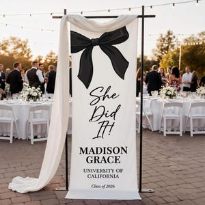 May include: A white banner with a large black bow illustration and the text "She Did It!" in elegant script. Below, it reads "MADISON GRACE, UNIVERSITY OF CALIFORNIA, Class of 2026." The banner is part of a graduation celebration.