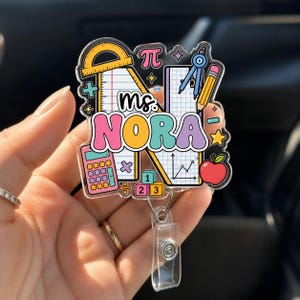 Peut inclure: Un porte-badge en acrylique transparent arborant une grande lettre N décorée d'illustrations sur le thème des mathématiques, dont un rapporteur, une calculatrice, un crayon, un compas, une pomme et des cubes. Le texte "Ms. NORA" est imprimé en lettres colorées, grasses et arrondies.