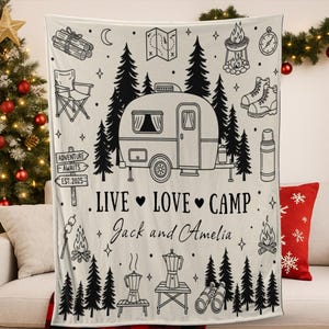 Puede incluir: Manta crema con diseño de camping en negro, con una caravana, árboles y equipo de acampada. El texto dice "LIVE LOVE CAMP" y nombres personalizados. Una manta acogedora para acampadas.