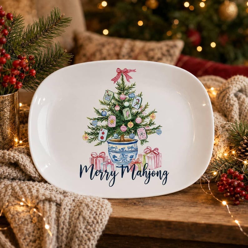 K&ouml;nnte beinhalten: Eine ovale wei&szlig;e Keramikplatte mit einer Aquarellillustration eines Weihnachtsbaums in einem blau-wei&szlig;en Topf, verziert mit Mahjong-Steinen und Kugeln, mit Geschenken darunter und dem blauen Schriftzug Merry Mahjong.