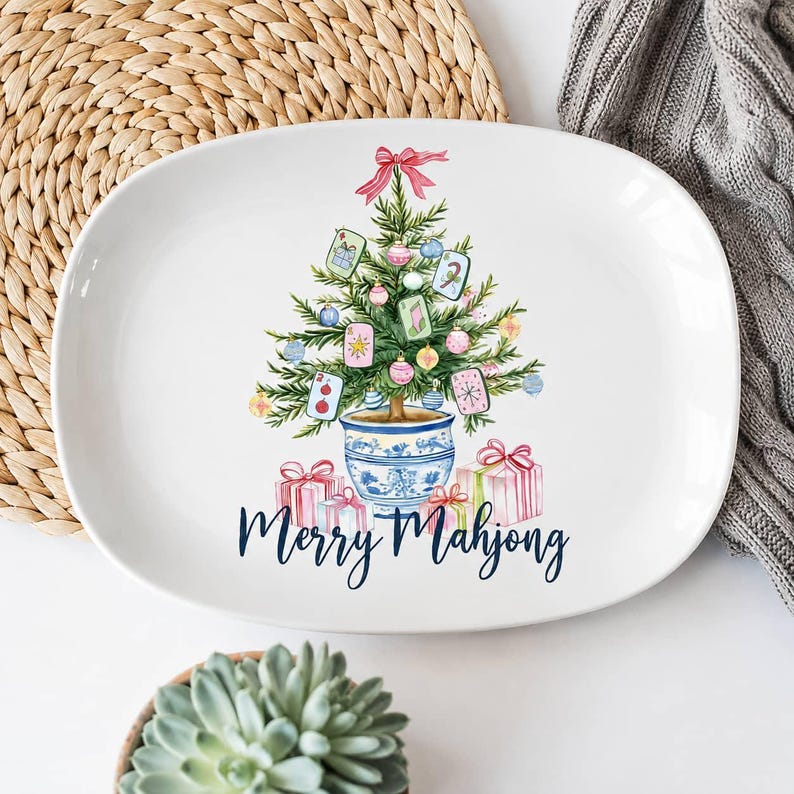 K&ouml;nnte beinhalten: Eine wei&szlig;e, ovale Keramikplatte mit einer Aquarellillustration eines Weihnachtsbaums in einem blau-wei&szlig;en Topf, verziert mit Mahjong-Steinen und Kugeln, sowie Geschenkboxen und dem Schriftzug Merry Mahjong in dunkelblauer Schrift.