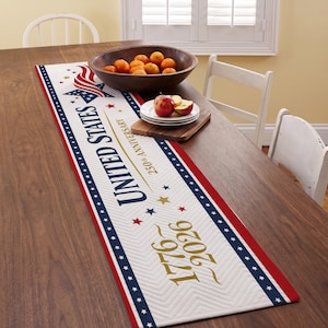 May include: A patriotic table runner with a white background, red and blue borders, and the text "UNITED STATES 1776 2026". The runner features stars and stripes, and is decorated with a wooden bowl of oranges and a plate of apples.