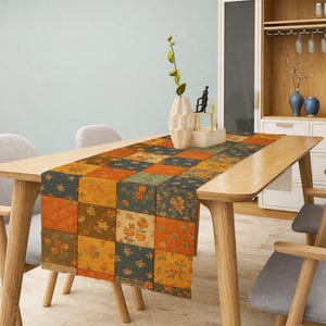 May include: A patchwork table runner with a floral pattern in shades of orange, yellow, and green. The runner is placed on a wooden table with gray chairs. A vase with a plant and decorative items are on the table.