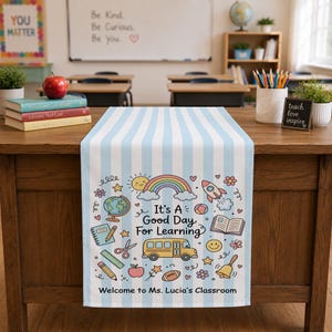Könnte beinhalten: Ein hellblau-weiß gestreifter Tischläufer mit bunten Illustrationen von einem Schulbus, Regenbogen, Globus, Büchern und Stiften. Der Text lautet "It's A Good Day For Learning" und "Welcome to Ms. Lucia's Classroom" in schwarzer Schrift.