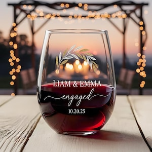 Op de afbeelding: Een stamloze wijnglas gevuld met rode wijn. Het glas is versierd met een gouden en wit bladontwerp en de woorden "Liam & Emma engaged 10.20.25" in elegant schrift. De achtergrond is een wazige buitenscène.